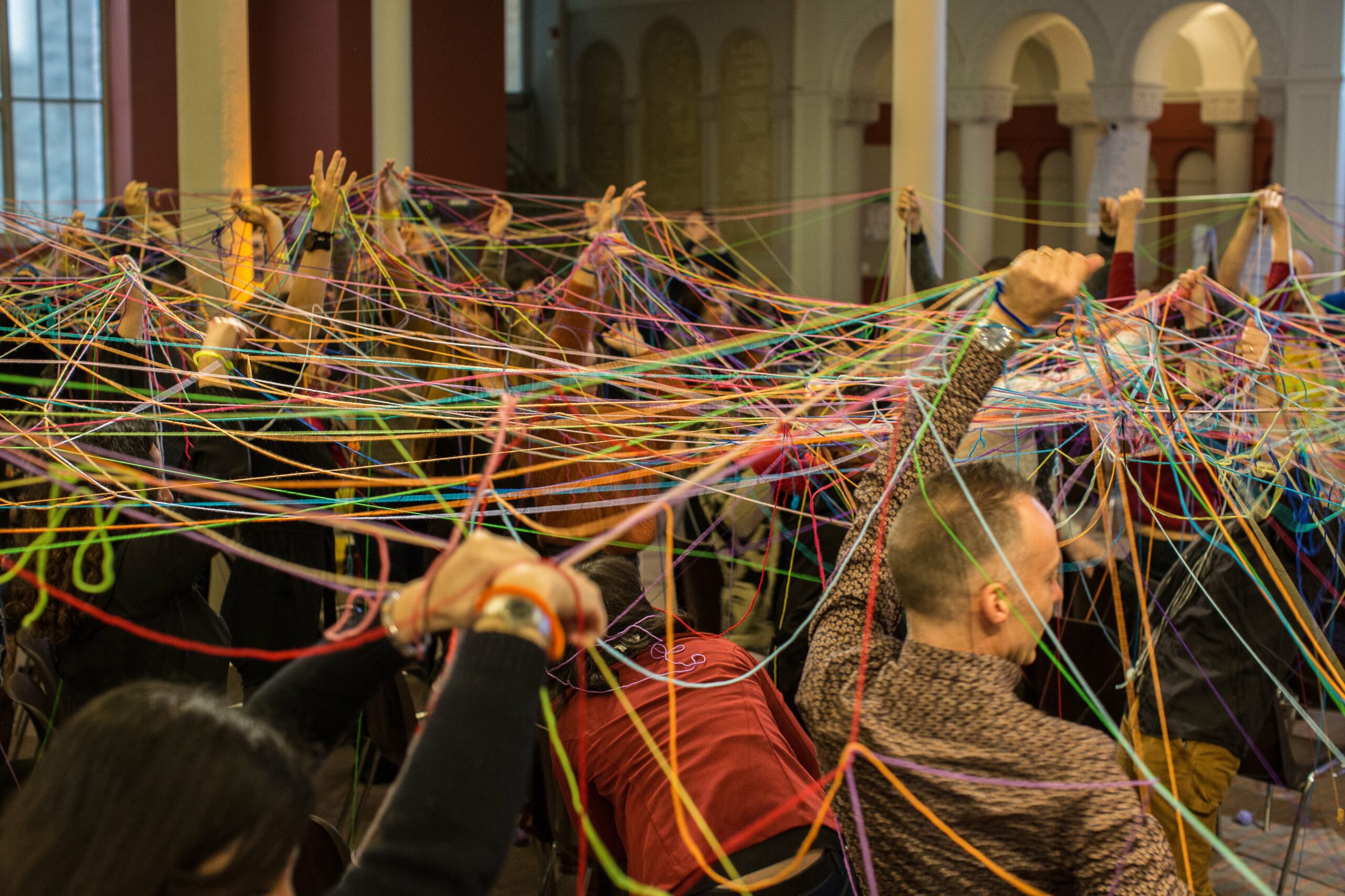 Un groupe de personnes sont reliés les uns aux autres par des fils de laine de toutes les couleurs qu'ils tiennent dans leurs mains, au-dessus de leur tête.