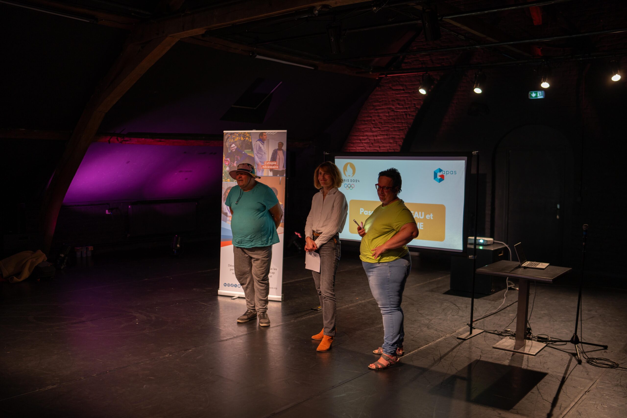 Sur une scène, trois personnes sourient, derrière elle, se trouvent un écran et un roll-up du Gapas.